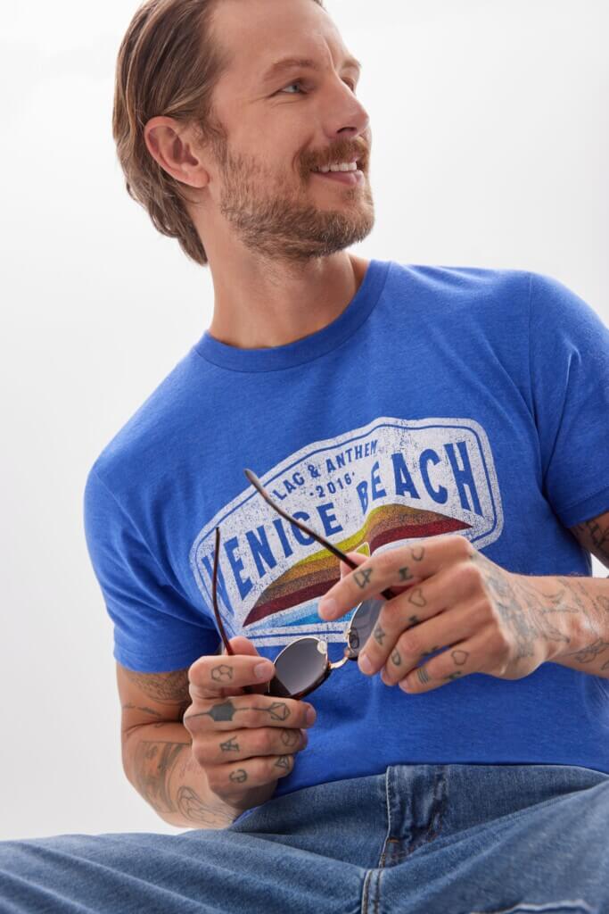 Man sitting and smiling holding sunglasses and wearing a blue T-shirt and jeans.