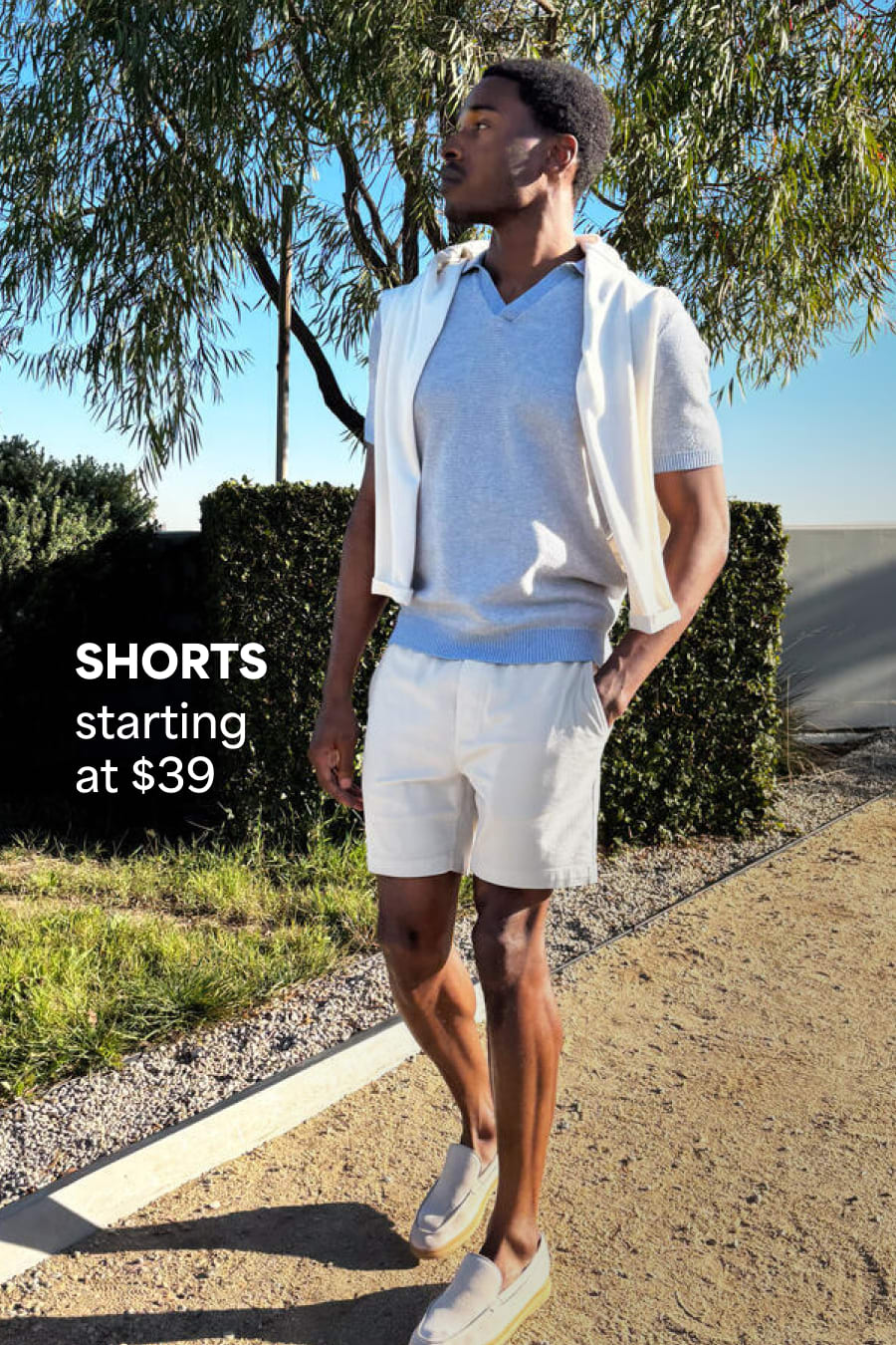 Model wearing Stitch Fix men’s clothes including white cardigan, short-sleeve blue polo and white shorts. Text reads: Shorts starting at $39.