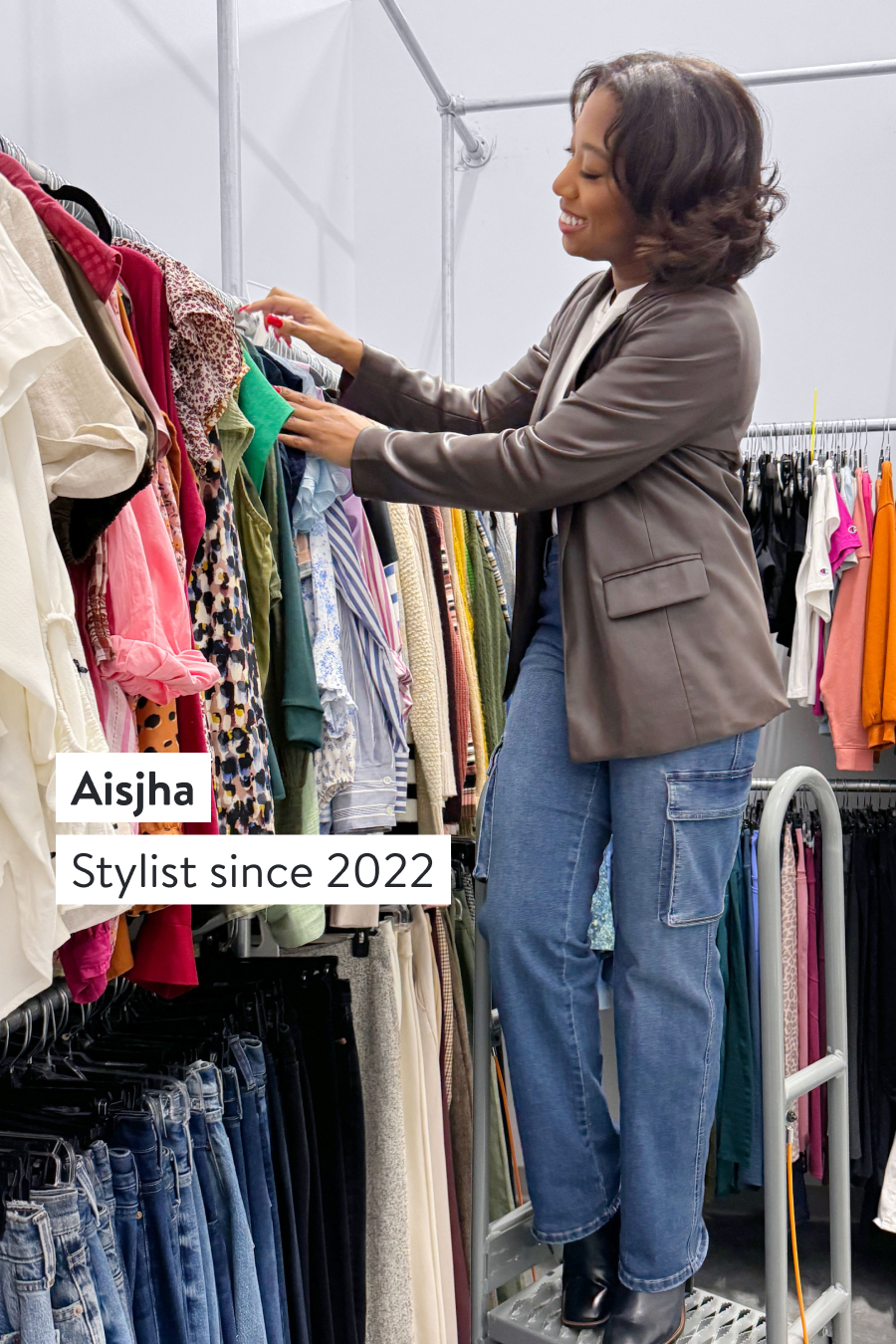 Aisjha, Stylist since 2022. Stitch Fix Stylist wearing a brown blazer stands on a ladder to select items including blouses, jeans, blazers and sweaters. 